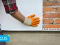 Installazione battiscopa per cappotto esterno: un operaio con guanti arancioni posiziona un pannello isolante bianco su una parete di mattoni, con un profilo metallico alla base.