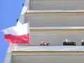 Prawidłowa flaga Polski powiewa na tle błękitnego nieba, obok balkonów z kwiatami.