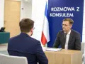Rozmowa z konsulem. Mężczyzna w okularach i garniturze siedzi przy biurku, obok flaga.