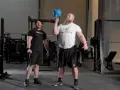 Man med skägg utför en kettlebell press med en blå kettlebell. En annan man står bredvid och tittar.