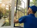 Man in blue shirt and cap performs window screen installation, preparing to fit the mesh into the frame.