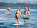 Eine Gruppe von Frauen genießt das Stand-Up-Paddling auf einem See. Sie stehen auf ihren Boards und paddeln durch das ruhige Wasser, während im Hintergrund Berge und Bäume zu sehen sind.