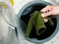 Main verte, petite, tenue dans une main, devant une machine à laver ouverte. Le coton rétrécit il au lavage ?