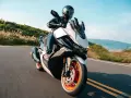 Kymco Xciting 400i dane techniczne: skuter z kierowcą na krętej drodze, z widokiem na góry i jezioro w tle.