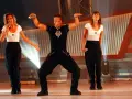 Drei Künstler in Schwarz-Weiß-Outfits performen auf einer Bühne. Die Frau links singt, der Mann in der Mitte tanzt, die Frau rechts hält ein Mikrofon. Ein energiegeladener Moment für **dance & electronic** Musik.