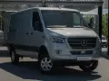 Srebrna budowa ciężarówki, model Mercedesa Sprintera, stoi na parkingu przed nowoczesnym budynkiem.