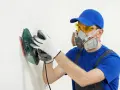 Pracownik w masce i okularach ochronnych wykonuje ręczne szlifowanie gładzi na białej ścianie za pomocą szlifierki.