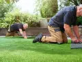 Dos hombres trabajan en un jardín, instalando césped artificial. Uno corta el borde mientras el otro lo alisa.