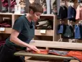 Mitarbeiterin pflegt orangefarbene Hose, damit sie für den Verkauf wie neu wachsen kann. Im Hintergrund sind Kleidung und Mützen ausgestellt.