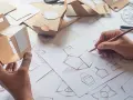 Hände skizzieren und falten Kartonmodelle für ein Produktdesign Studium. Verschiedene Verpackungsentwürfe liegen auf dem Tisch.