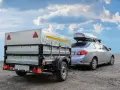 Un'auto argento con box sul tetto traina un rimorchio chiuso. La differenza tra carrello appendice e rimorchio è chiara: questo è un rimorchio.