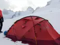 Alpinista w czerwonej kurtce stoi obok namiotu na tle ośnieżonych szczytów. W oddali widać groźne góry, gdzie mogą wystąpić lawiny w Alpach.