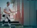 Man i arbetskläder använder en lång roller för att måla huset. En hink med färg står på gräset.