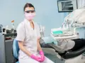 Asystentka stomatologiczna w masce i okularach ochronnych siedzi w gabinecie.