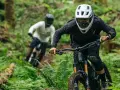 Riders in helmets and protective gear shredding a forest trail. This image shows what to wear mountain biking: full-face helmets, goggles, long sleeves, and knee pads.