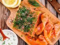 En bit gravad lax, generöst toppad med dill och flingsalt, ligger på en träskärbräda bredvid skivor av lax och en skål med sås.
