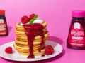 Ein Stapel zuckerfreie pancakes mit Himbeersauce und frischen Beeren. Daneben Flaschen mit Erdbeer- und Himbeer-Topping.