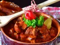 Aromatyczny, tradycyjny gulasz węgierski w glinianej misie, udekorowany papryczką i natką. Obok pieczywo i drewniana łyżka z pieprzem.