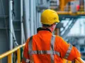 Elektryk w żółtym kasku i pomarańczowej kamizelce odblaskowej na tle przemysłowej konstrukcji.