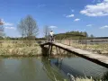 Osoba z rowerem na drewnianym mostku nad rzeką. Piękny dzień na odkrywanie tras rowerowych łódzkiego.