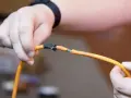 Dłonie trzymające pomarańczowy kabel z odsłoniętymi żyłkami, prowizorycznie połączone taśmą. Pokazuje, jak połączyć kable bez lutowania.