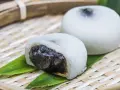 Dwa białe mochi, jedno przekrojone, ukazujące ciemne nadzienie. Podane na bambusowej tacce z liśćmi.