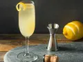 Elegancki drink z szampanem, ozdobiony skórką cytryny, obok kieliszka stoi miarka barmańska, cytryna i korek od szampana.
