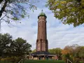 Der Aussichtsturm im Volkspark Jungfernheide ragt mit seinem roten Backstein und grünen Kuppeldach in den Himmel. Herbstlaub umrahmt die Szene.