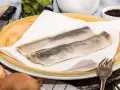 Zwei Matjesfilets auf Küchenpapier, bereit für eine gesunde Mahlzeit. Zwiebeln und Pfefferkörner deuten auf die Zubereitung hin.