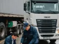 Dwóch mężczyzn przy ciężarówce sprawdza dokumenty. Na pierwszym planie widać fragmenty naczepy i opon. Jeden z nich trzyma w ręku kartę przewozową.