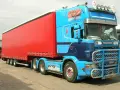 Naczepa jumbo w jaskrawym kolorze czerwonym, ciągnięta przez niebieską Scanię z malowidłami.