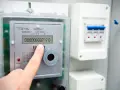 Palec wskazuje na cyfrowy licznik energii elektrycznej, pokazujący odczyt 00000000717.0. Widoczne są symbole na liczniku energii elektrycznej.