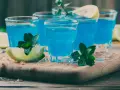 Kilka shotów z drinkiem blue curacao, ozdobionych limonką i miętą, na drewnianej desce z lodem.