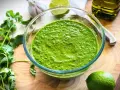 Frisch zubereitetes mojo verde in einer Glasschüssel, umgeben von Koriander, Knoblauch, Limetten und Olivenöl.