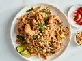 Pyszny Pad Thai przepis: makaron ryżowy z krewetkami, jajkiem, orzeszkami ziemnymi i szczypiorkiem. Obok chili i orzeszki.