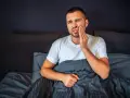 Mężczyzna w łóżku cierpi z powodu tragicznego bólu zęba, trzymając się za policzek.