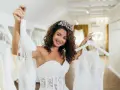 Uśmiechnięta panna młoda w koronie wybiera suknię ślubną. Zastanawia się, ile kosztuje wypożyczenie sukni ślubnej, porównując dwie piękne kreacje.
