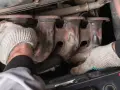 Mechanic's gloved hands work on a rusty exhaust manifold, possibly to fix an engine ticking noise.