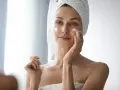 Femme appliquant de la crème sur son visage pour lutter contre la xerose cutanée. Elle a une serviette sur la tête.