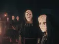 Miembros del culto de Kosmos, con túnicas oscuras y máscaras blancas, se reúnen ante un objeto piramidal brillante.