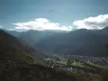 Vue aérienne d'une vallée verdoyante avec un village niché, entouré de montagnes majestueuses. Un planeur plane dans le ciel bleu, rappelant les paysages de Toulouse et ses environs.