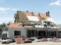 Ekipa budowlana pracuje nad remontem dachu domu. Widać rusztowania, drabiny i materiały budowlane.
