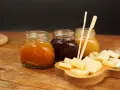 Tre vasetti di marmellata, uno arancione, uno scuro e uno giallo, accompagnano un tagliere di formaggio a cubetti, pronto per essere gustato.
