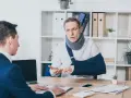 Mężczyzna w kołnierzu ortopedycznym i temblaku rozmawia z drugim mężczyzną przy biurku.