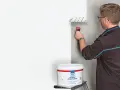 Mann streicht mit einer Farbrolle eine Wand weiß. Der Eimer mit der "Profi Silikat Wandfarbe" steht daneben. Perfekt für das Streichen bei Auszug.