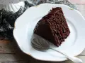 Una fetta di torta al cioccolato morbida e umida, ricoperta di glassa lucida, su un piatto bianco con un cucchiaino.