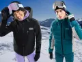 Coppia con abbigliamento da sci, giacche hardshell e maschere da neve, su sfondo montuoso innevato.