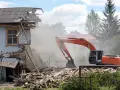 Koparka burzy zniszczony budynek, unosząc tumany kurzu. W tle inne domy i drzewa. Rozbiórka budynku formalności w toku.