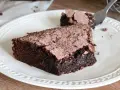 Una fetta di torta al cioccolato fondente, cosparsa di cacao, su un piatto bianco. Un pezzetto è stato staccato con una forchetta.
