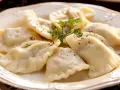 Talerz pełen idealnych, gotowanych pierogów, posypanych pieprzem i udekorowanych natką pietruszki.